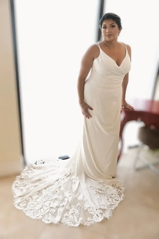 A woman in a white wedding dress with lace detailing stands near a large window, posing elegantly.