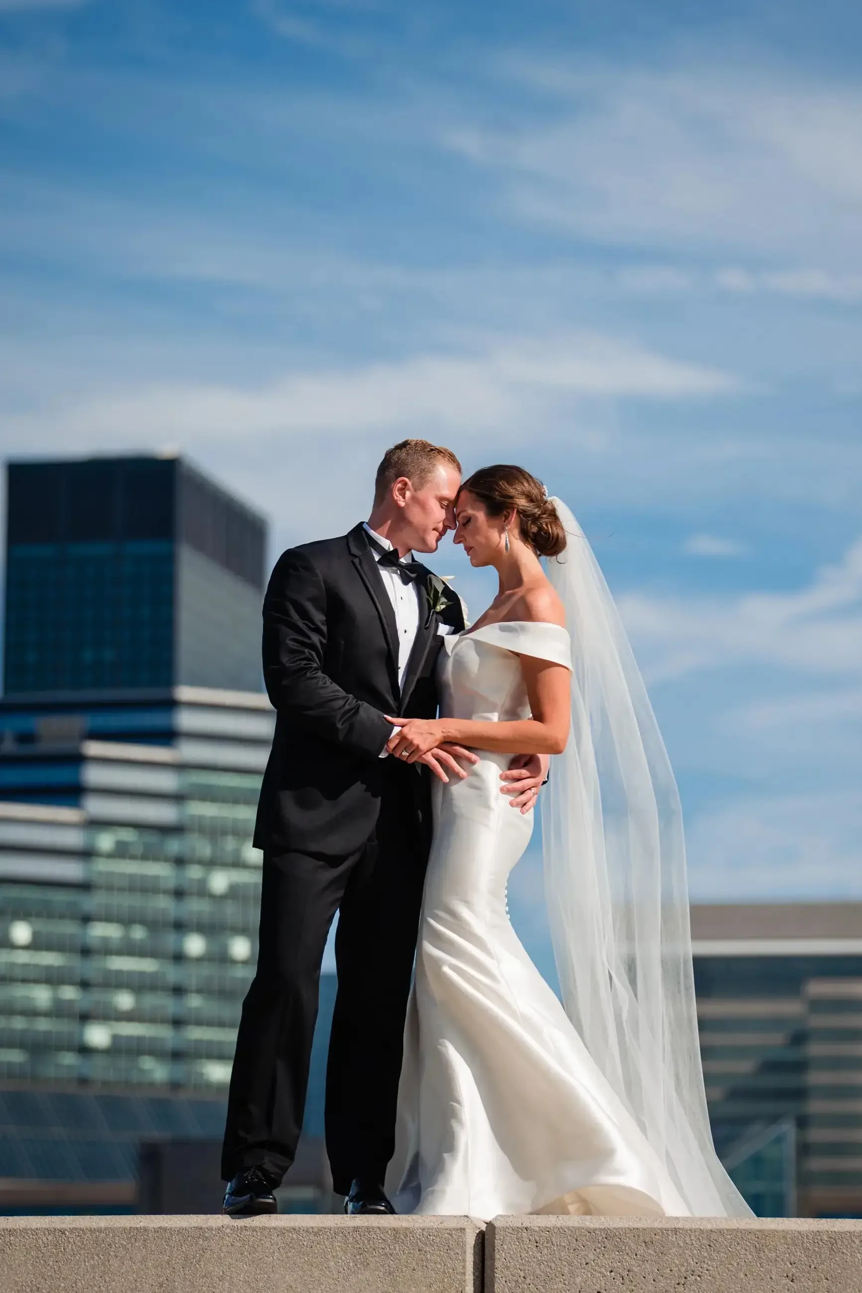 A bride and groom lean in towards each other in front of a cityscape on their wedding day