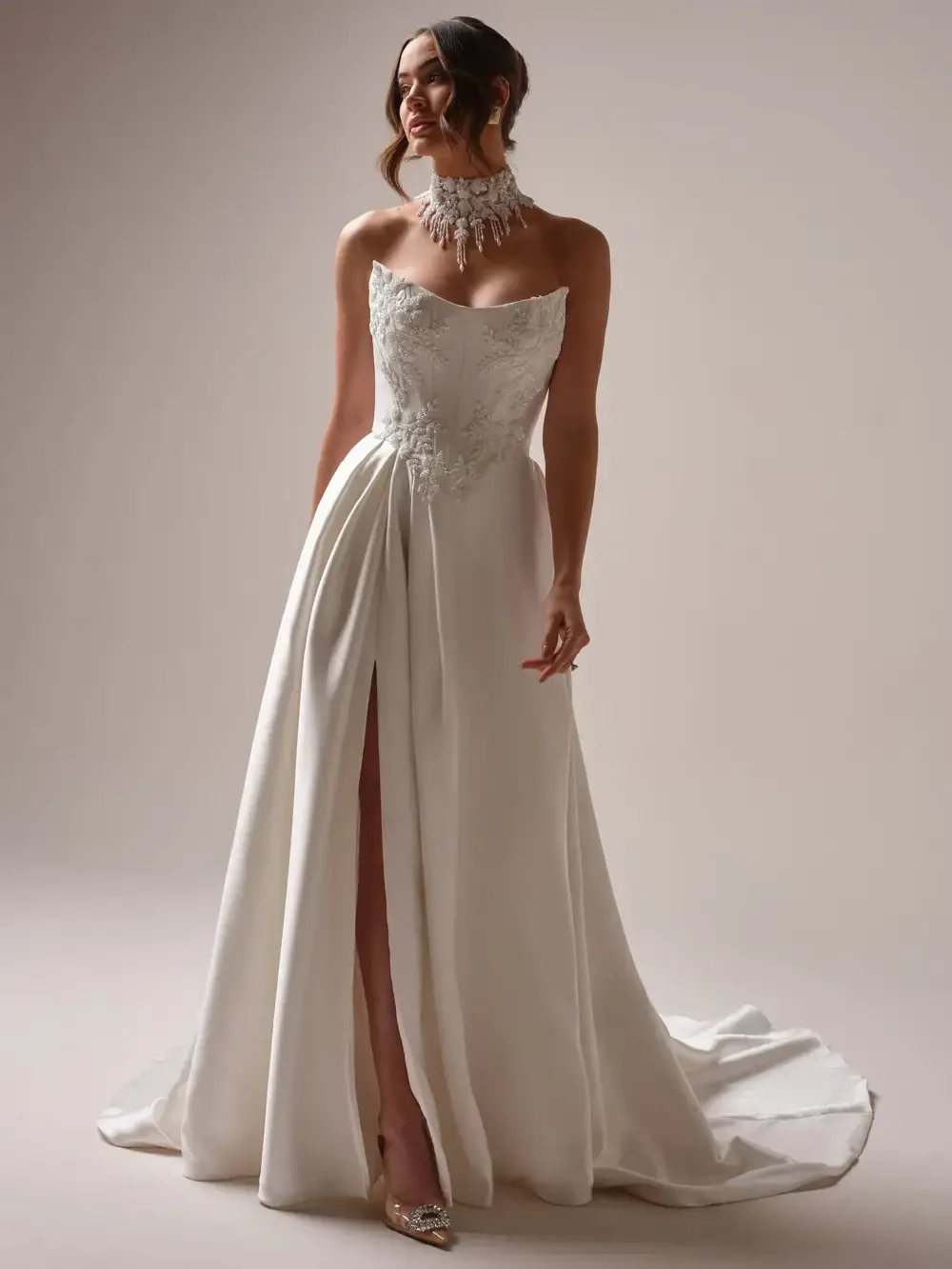 A bride wearing a strapless white wedding gown with intricate detailing and a high slit, accessorized with a sparkling necklace.