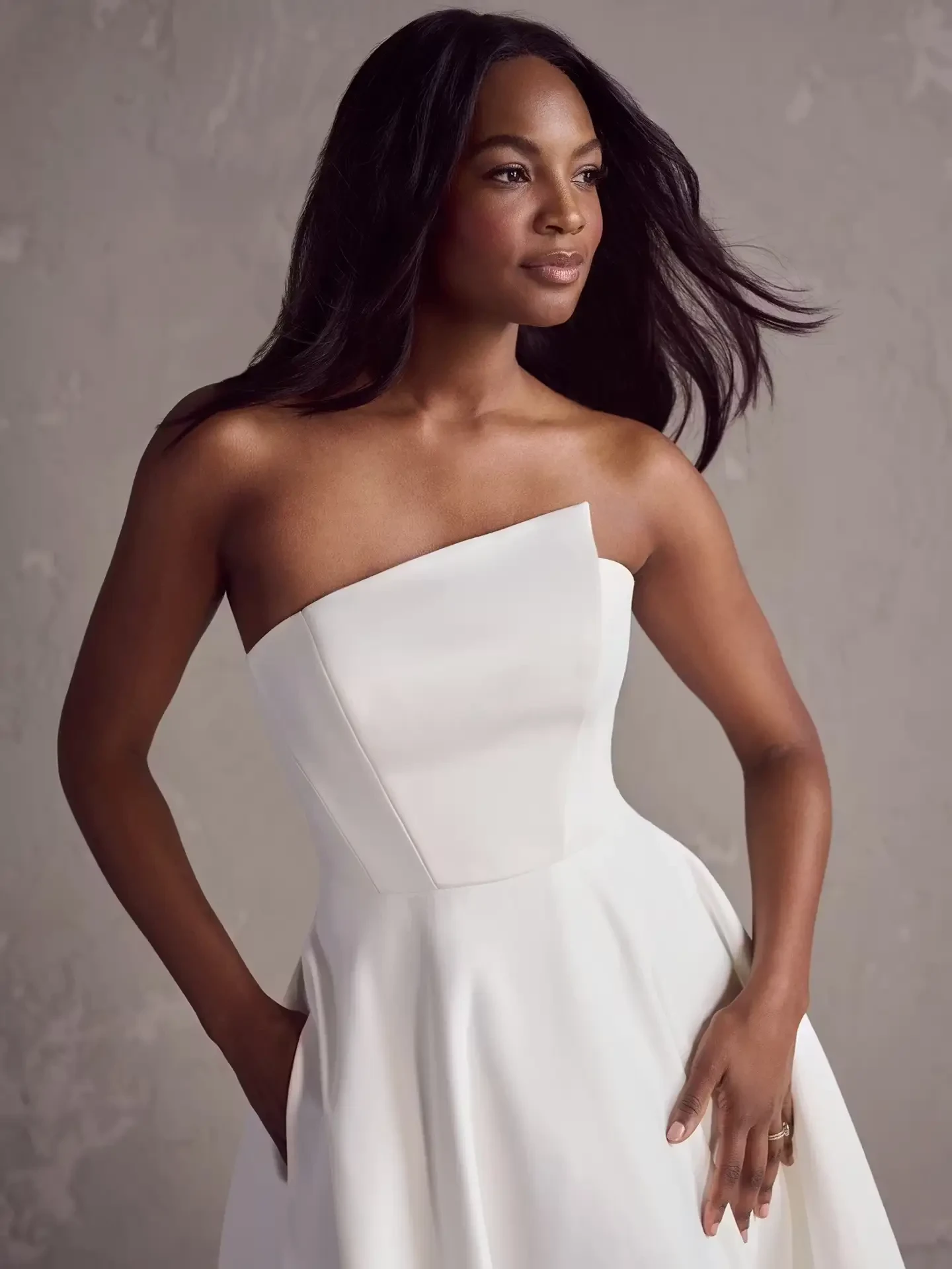 A woman wearing a strapless white dress, posing gracefully with one hand on her hip and the other relaxed by her side, against a neutral background.