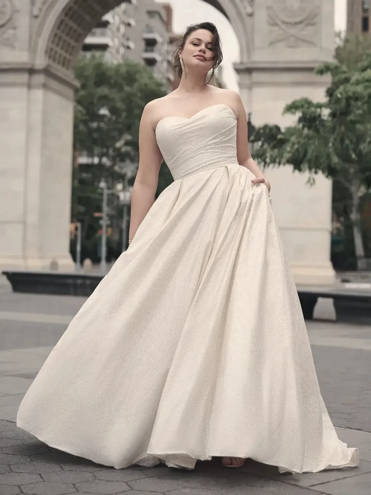 Woman in an elegant, strapless white gown poses confidently outdoors near an archway and trees, exuding grace and sophistication.