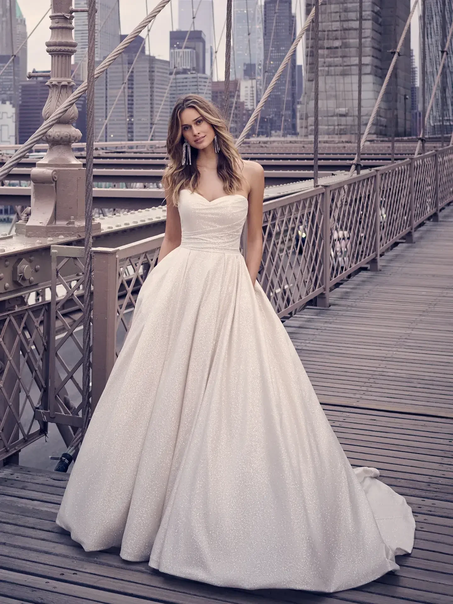 A woman in a strapless white wedding gown stands on a bridge with city skyscrapers in the background. The scene conveys elegance and romance.