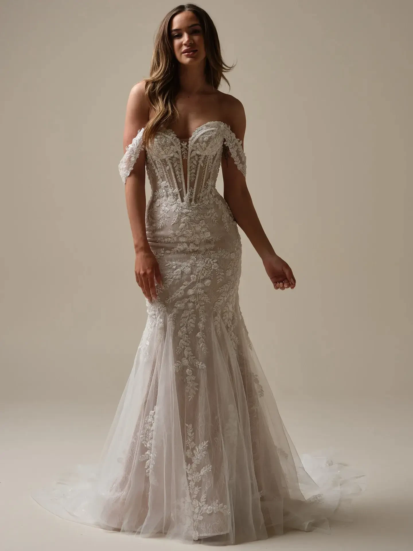 A woman is wearing an elegant, strapless wedding dress with lace details and a fitted silhouette that flares at the bottom. She has long hair and is standing confidently against a neutral background.
