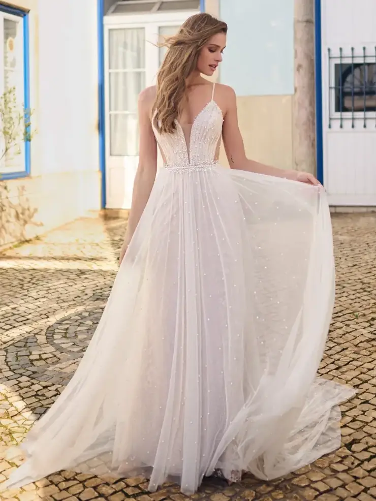 A woman in a white, flowing dress with delicate beading and straps, standing on a cobblestone path.