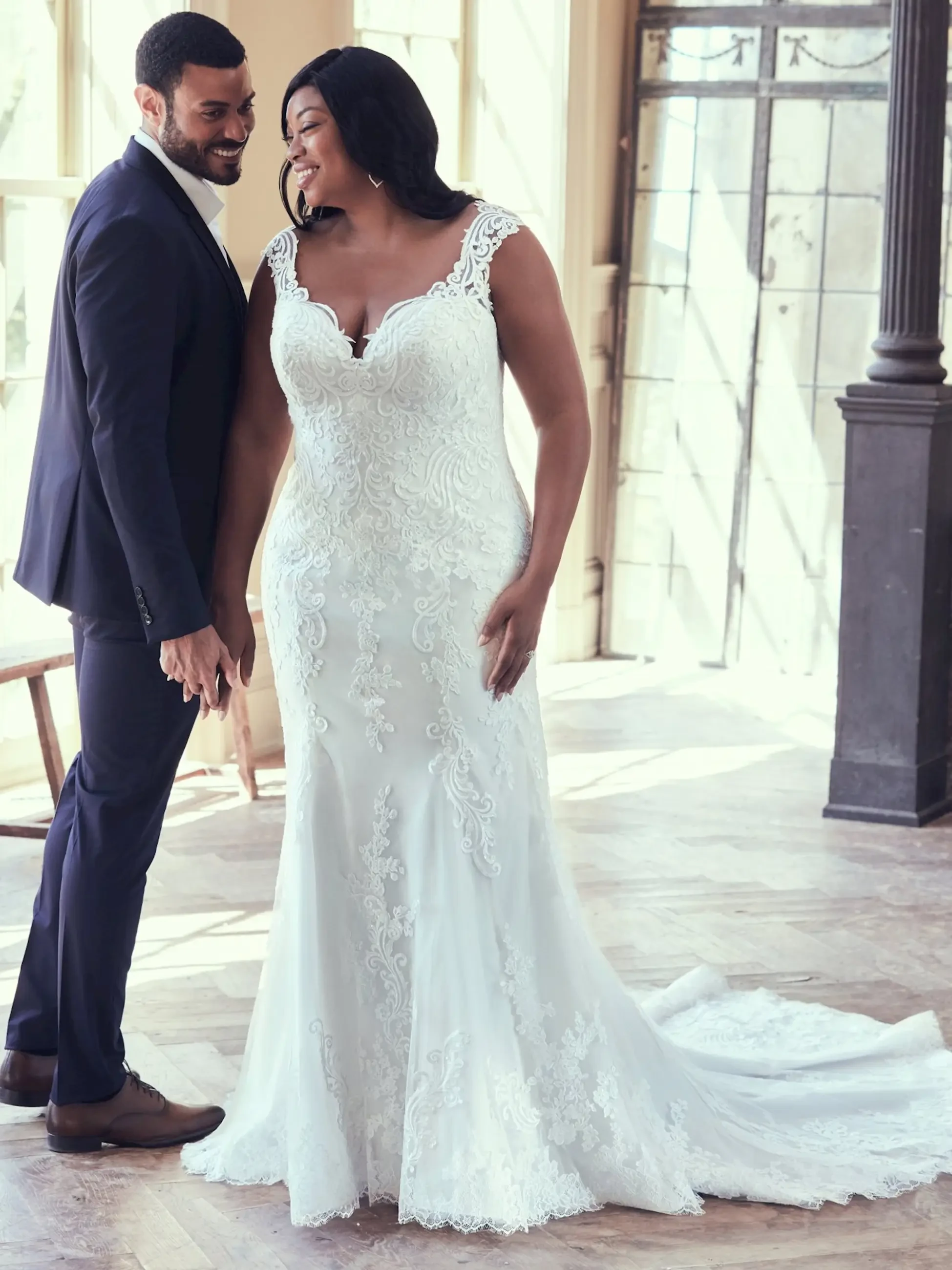 A smiling bride in an elegant lace wedding gown and groom in a dark suit stand closely, holding hands in a sunlit room, conveying joy and romance.