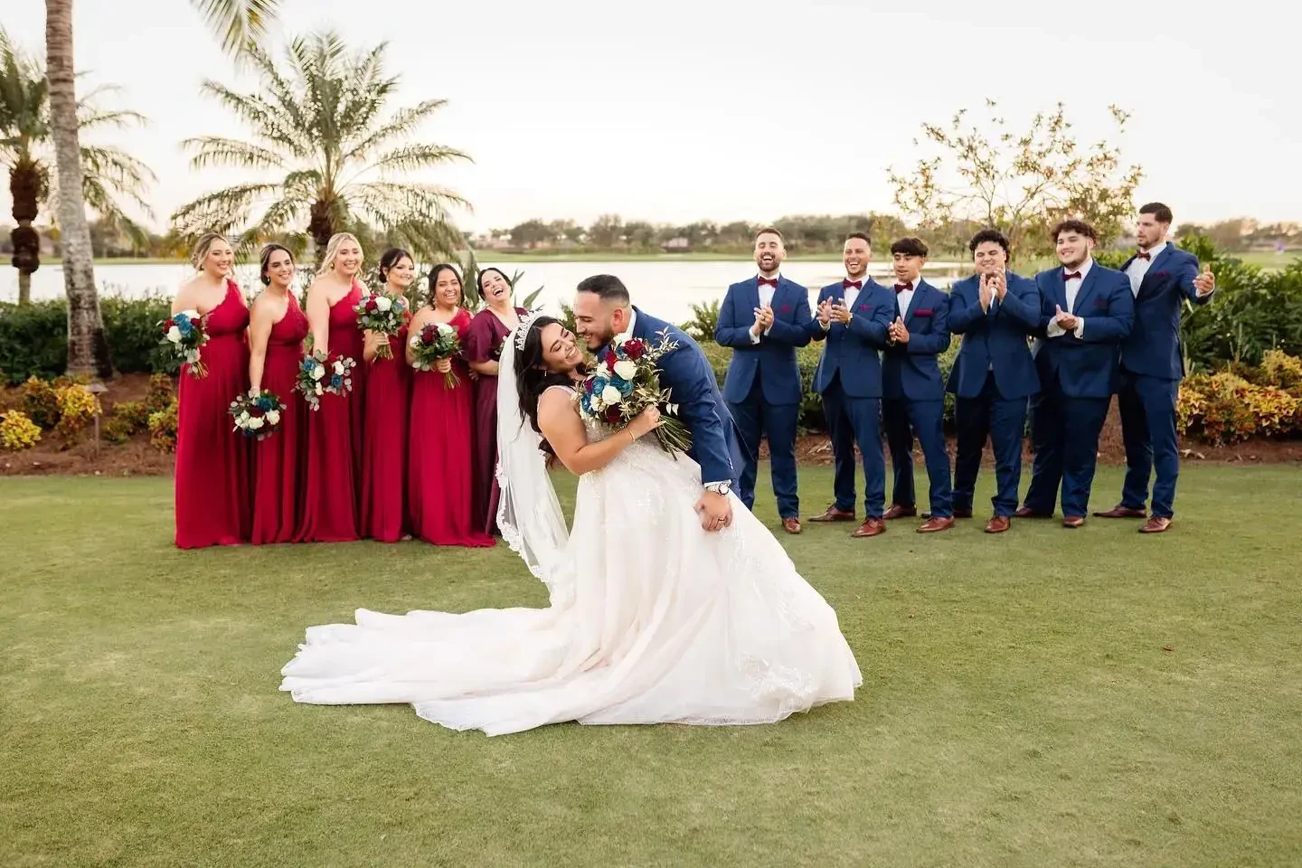 A groom dips his bride for a kiss while his groomsmen and her bridesmaids smile behind them in red gowns and navy blue suits