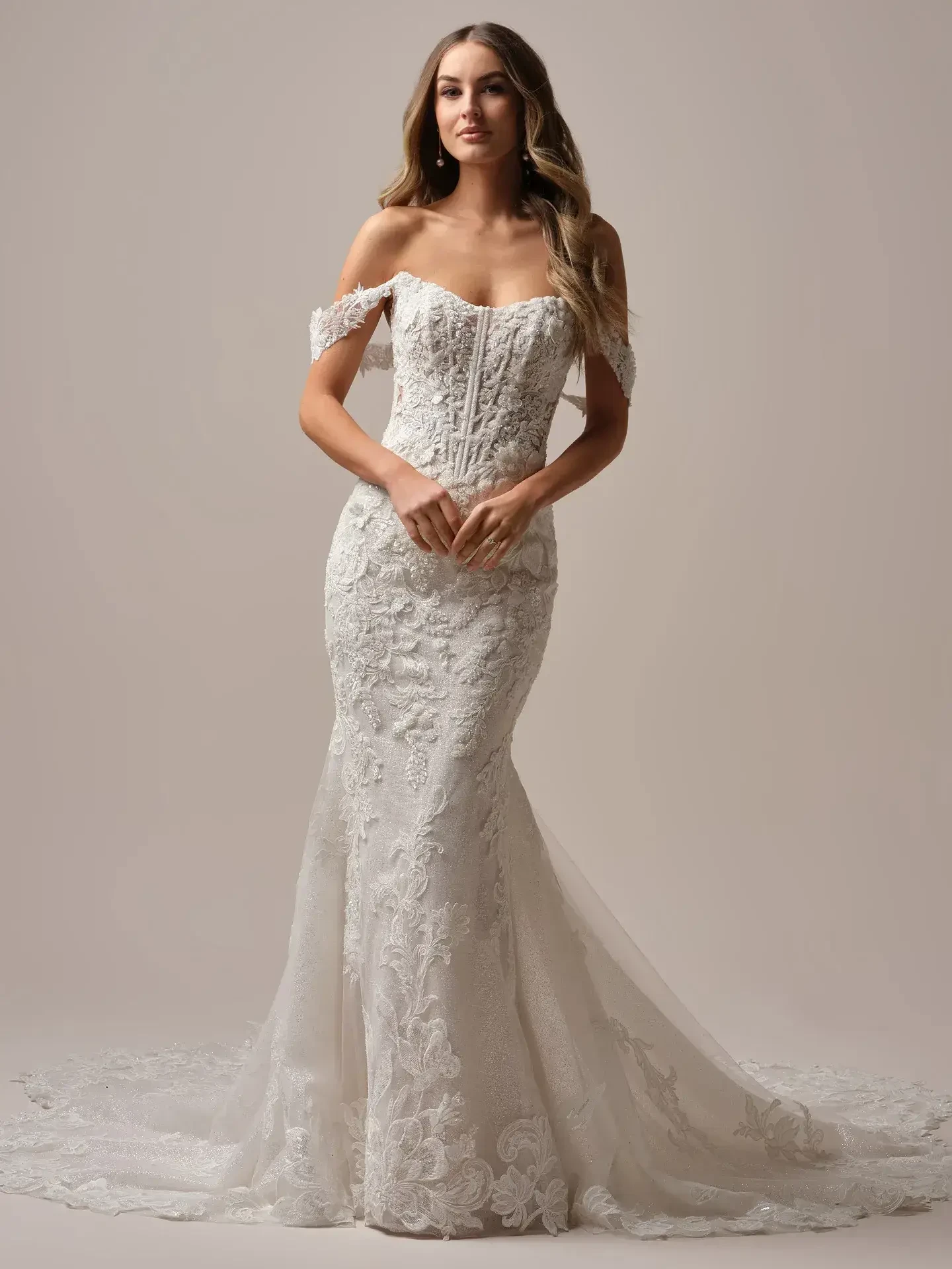 A woman wearing an elegant, off-the-shoulder lace wedding gown stands on a plain background. Her long, wavy hair and serene expression convey grace.