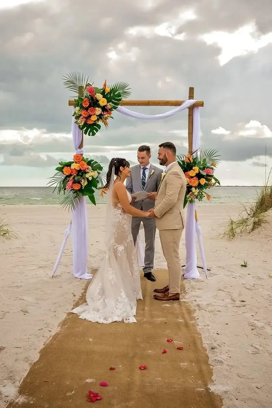 A bride and groom get married on a beach with rose petals scattered on the aisle and a beautiful arch behind them with orange and yellow flowers