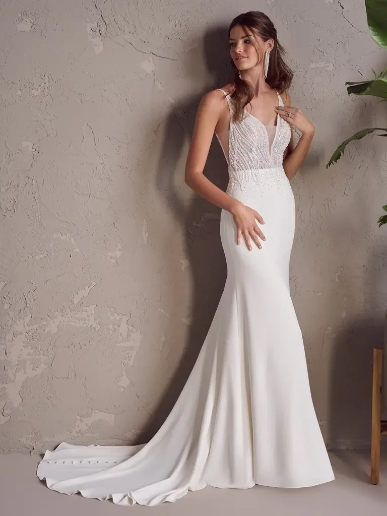 A model is wearing a fitted white wedding dress with a lace bodice and a flowing train, posing against a textured wall with plants nearby.