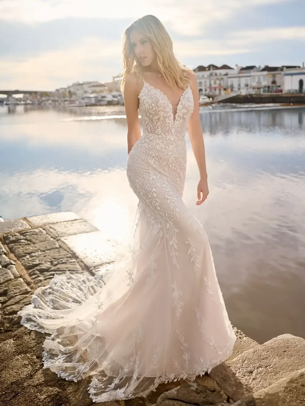 A woman in a wedding dress stands by a body of water, with a picturesque background of buildings and a bridge under a partly cloudy sky. The dress is form-fitting and features intricate lace detailing.