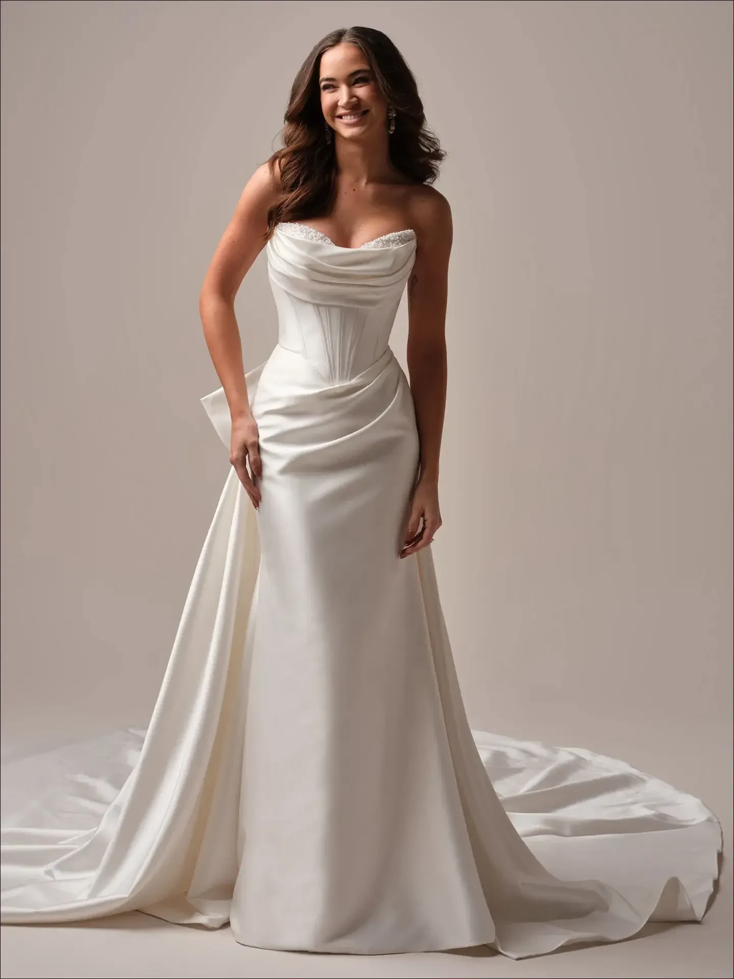 A woman in a strapless, satin wedding dress with a long train smiles softly. The dress features a fitted bodice and draped fabric, conveying elegance and joy.