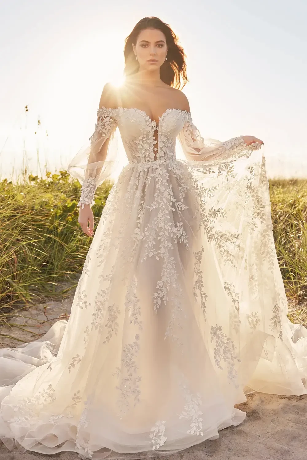 A woman in an elegant, off-shoulder wedding dress with floral lace details, standing on a beach during sunset.