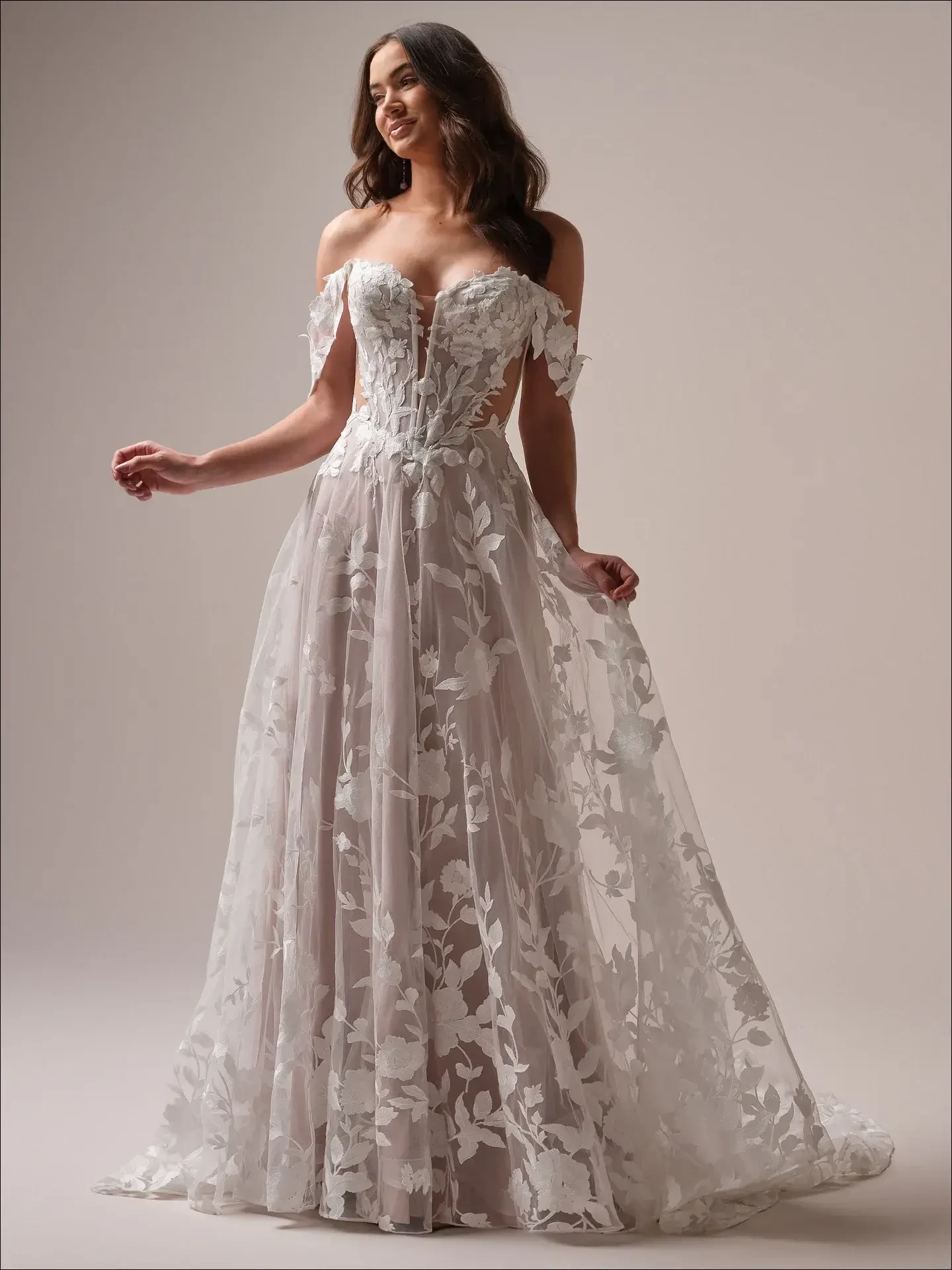 A woman in a flowing, off-shoulder wedding gown adorned with floral lace stands smiling. The elegant dress conveys a romantic, graceful tone.