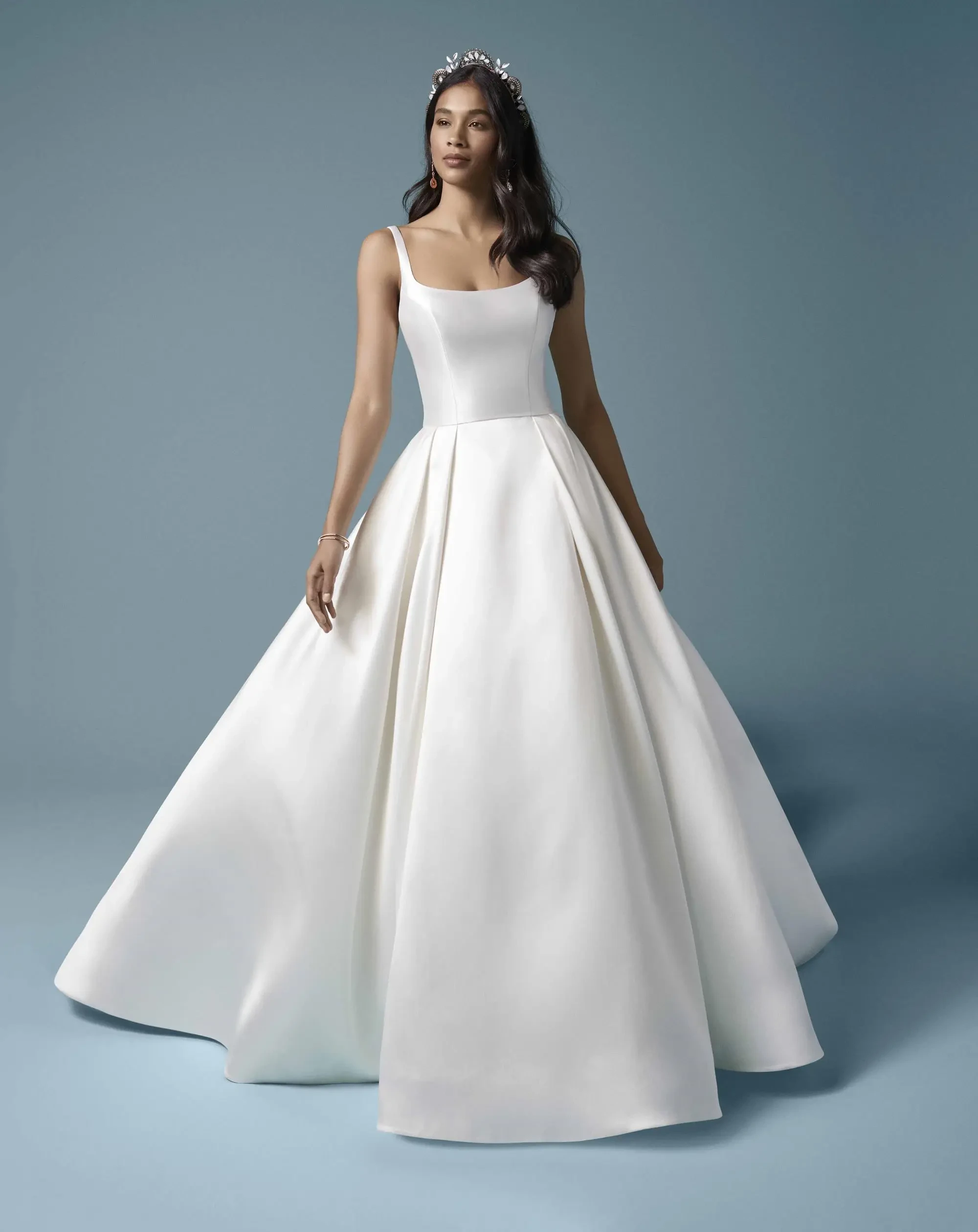 A model wearing an elegant white wedding dress with a fitted bodice and a flared skirt, posed against a soft blue background.