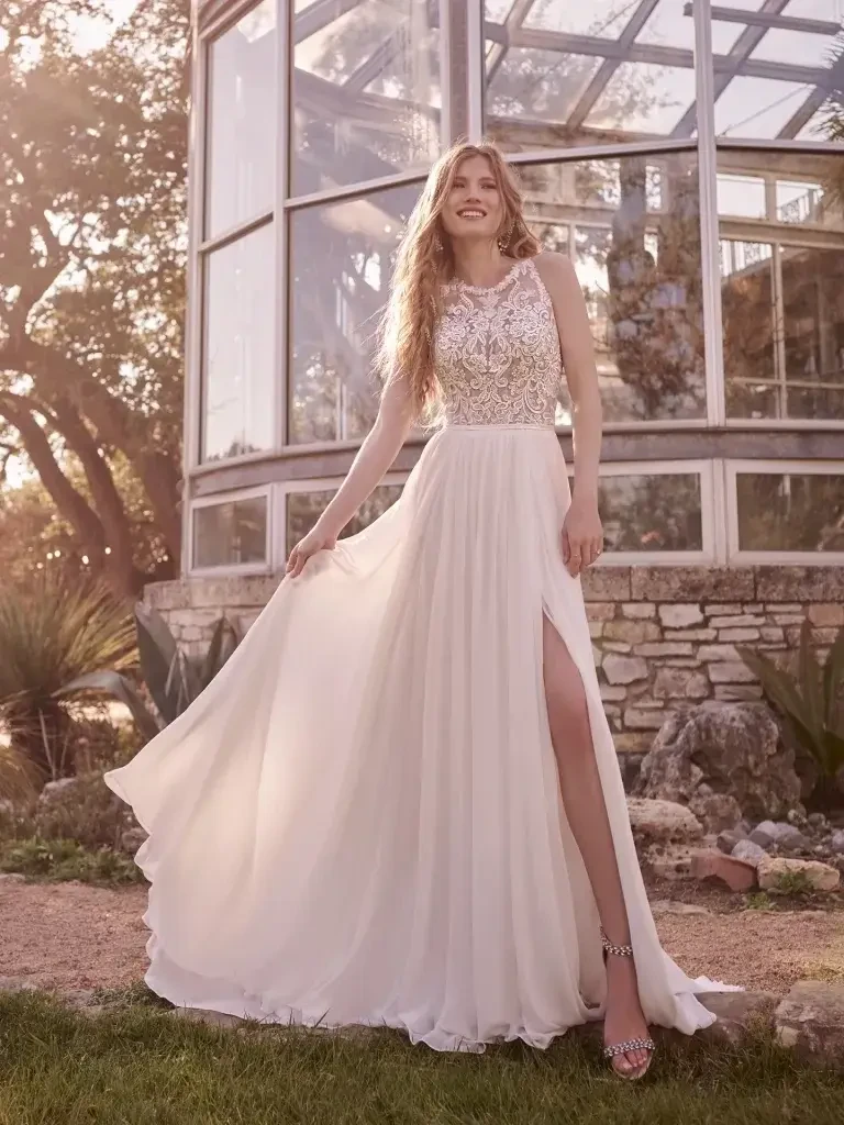 A woman in a flowing white dress with a beaded bodice stands outside near a glass greenhouse, smiling and holding the skirt of her dress.