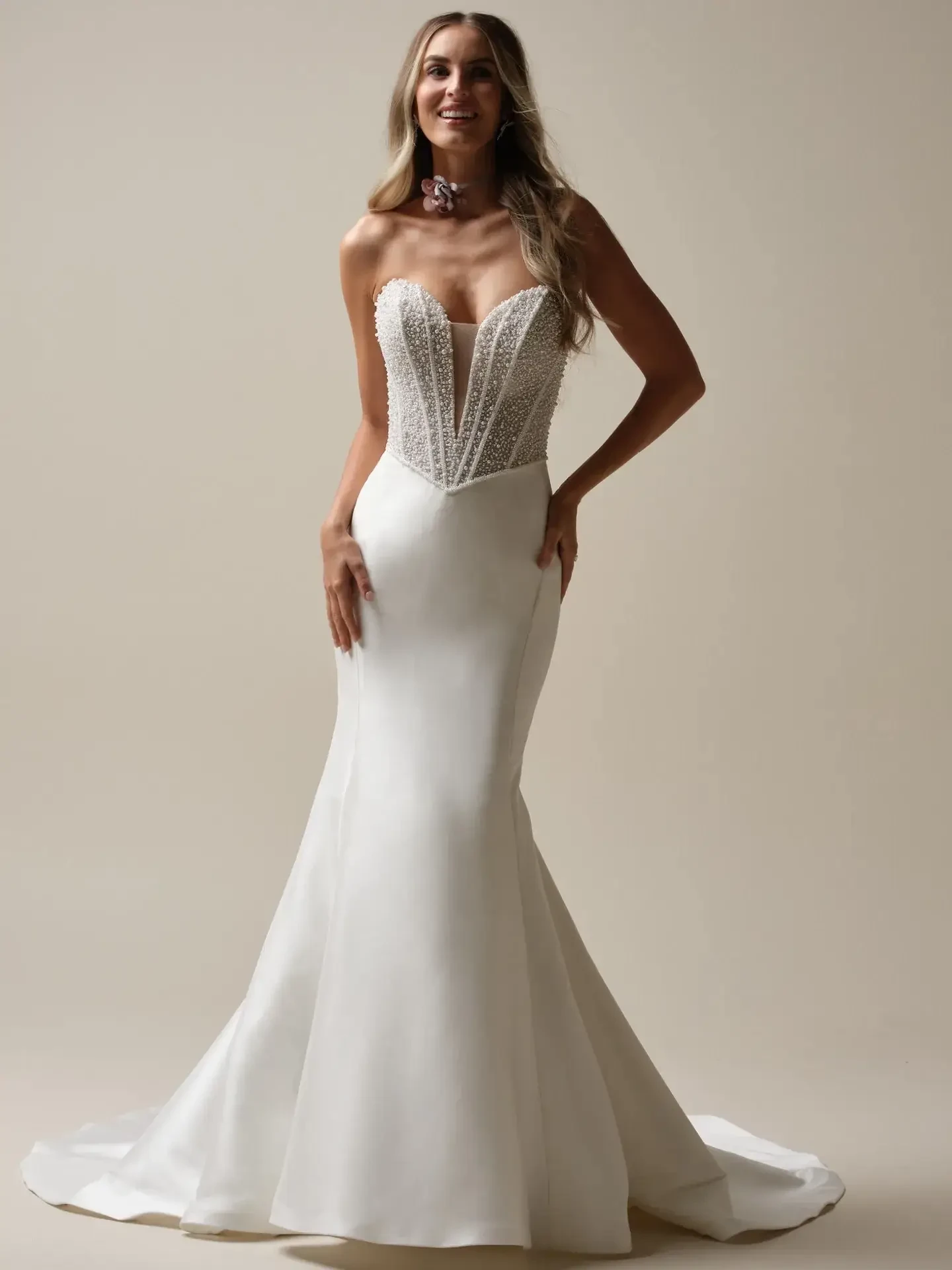 A woman wearing a fitted white strapless gown with intricate beading and a mermaid silhouette, standing against a neutral background.
