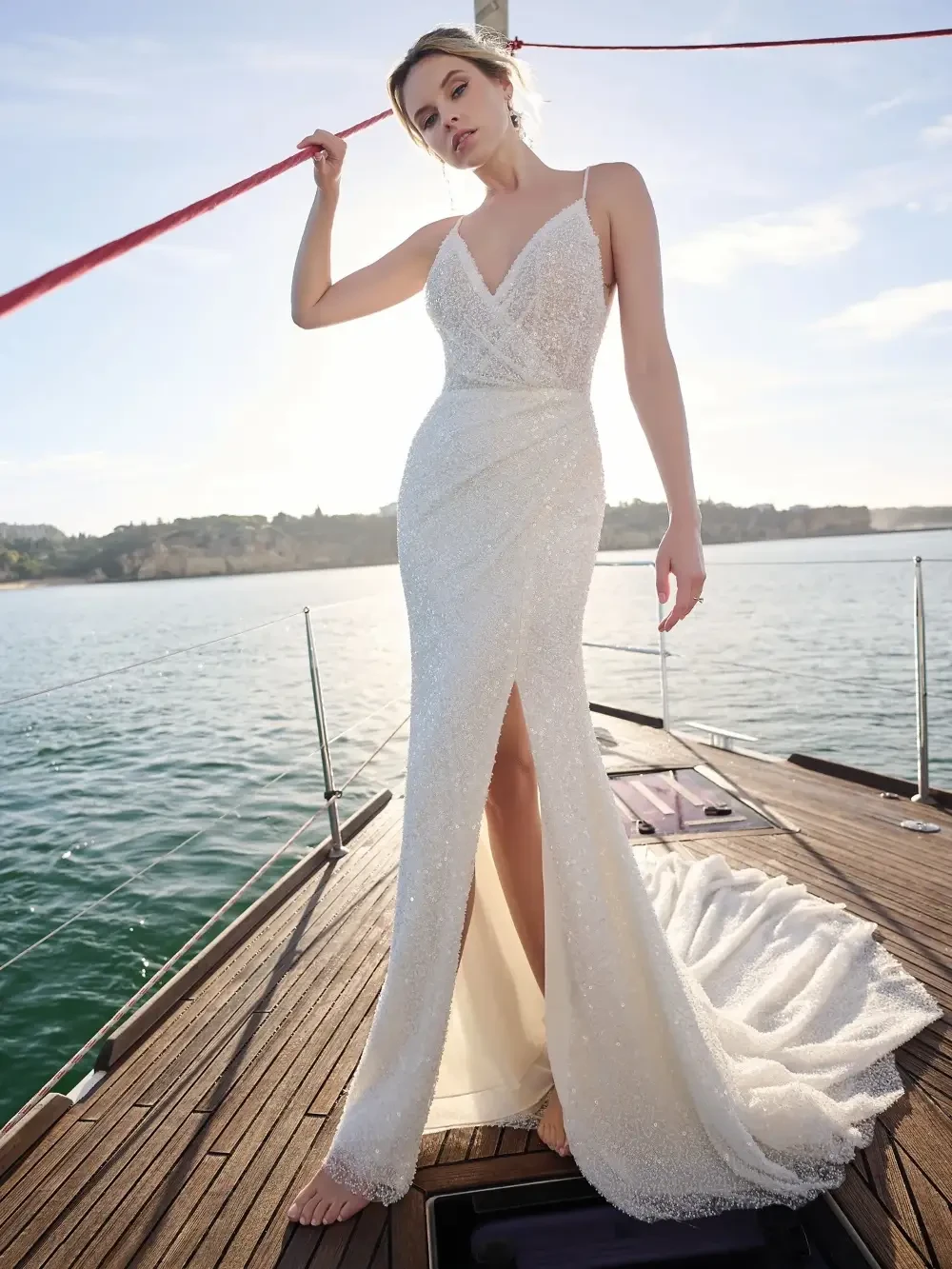 A model wearing a stunning white evening gown with a deep V-neckline and a thigh-high slit, standing on a boat with water in the background.