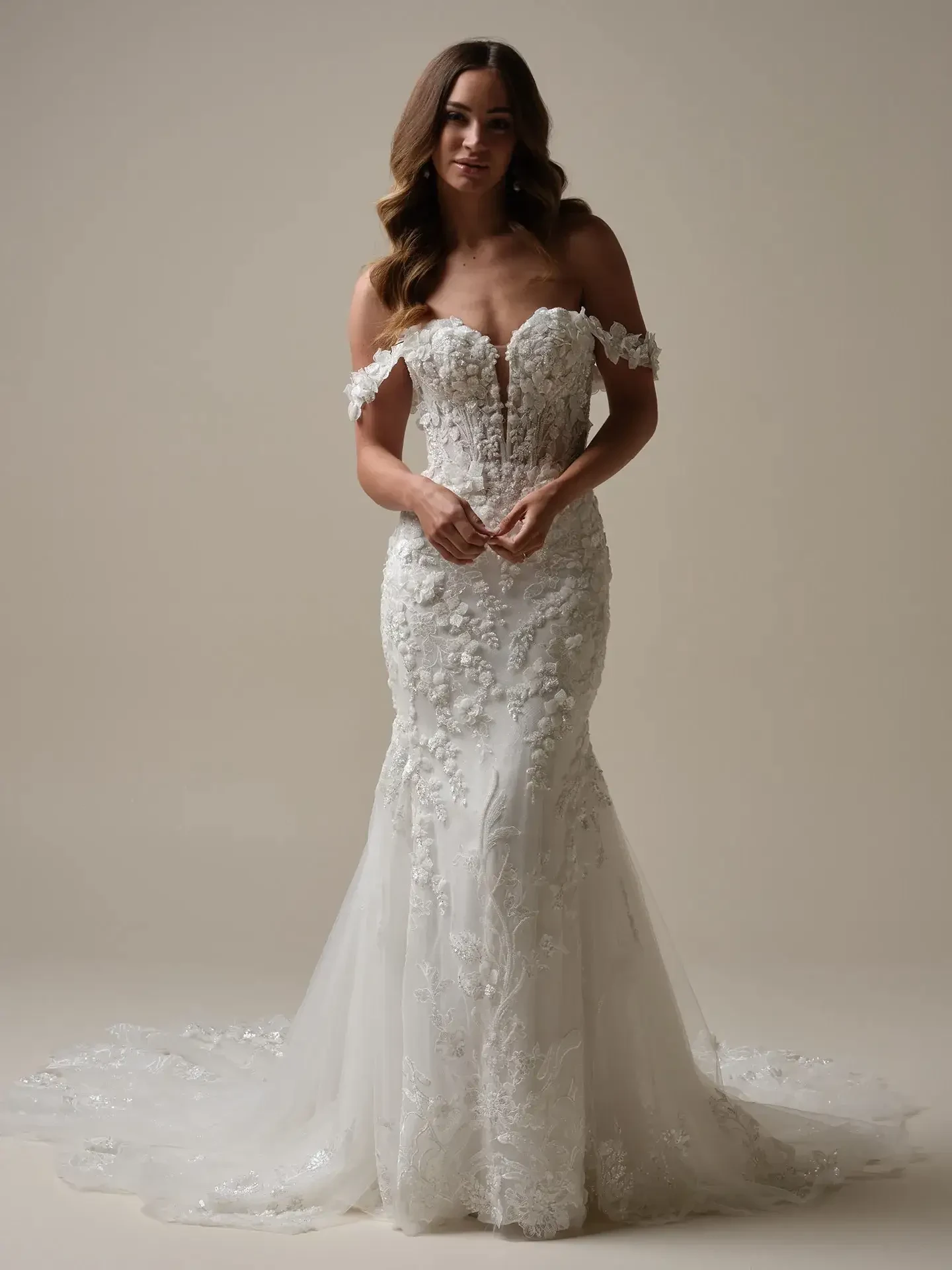 A woman in an elegant, off-the-shoulder white wedding dress with floral lace details stands against a neutral background, conveying a serene and graceful mood.